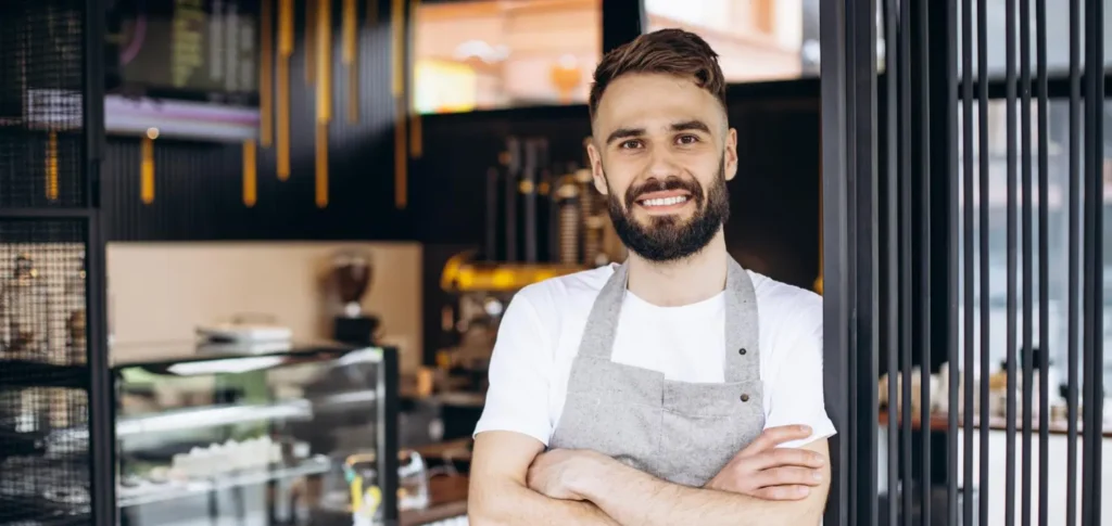 Restaurateur souriant en tablier, bras croisés, à l'entrée de son café-restaurant