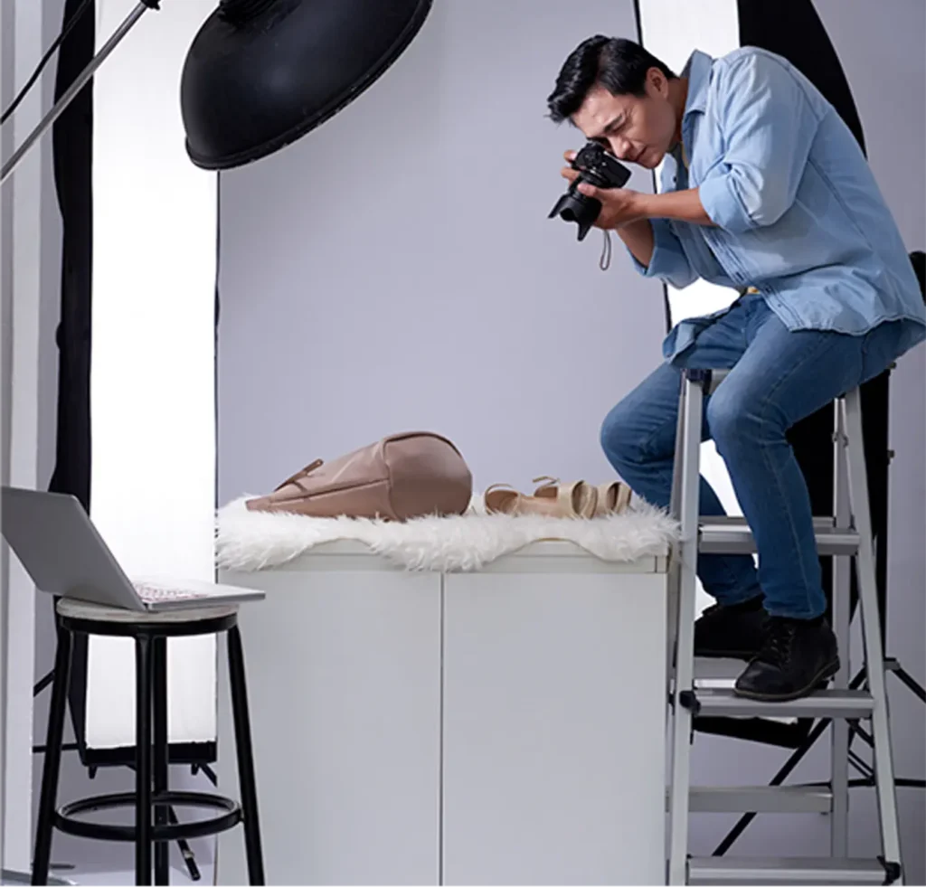 Photographe artiste-auteur réalisant un shooting produit en studio, assis sur un escabeau derrière son appareil