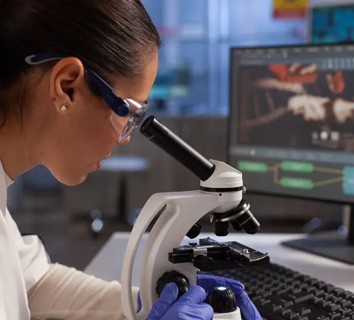 Chercheuse en blouse et lunettes de protection observant un échantillon au microscope dans un laboratoire